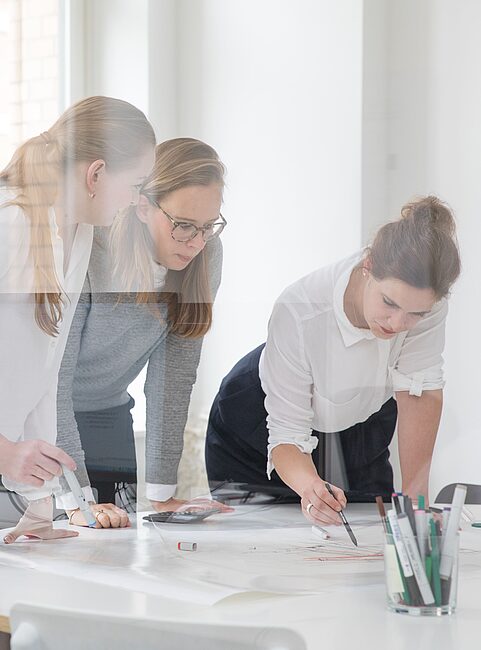 Drei Frauen stehen an einem Tisch mit Papier und Stiften in einem hellen Büro, arbeiten gemeinsam an einem Projekt.