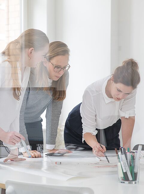 Drei Frauen stehen an einem Tisch mit Papier und Stiften in einem hellen Büro, arbeiten gemeinsam an einem Projekt.