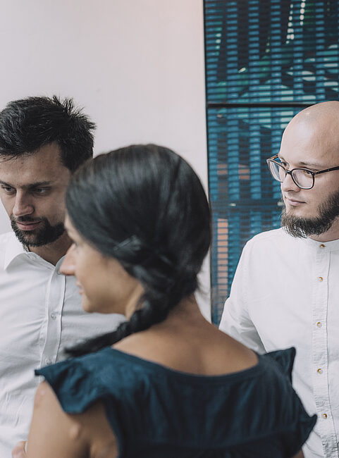 Vier Personen in weißen Hemden stehen zusammen, eine Person trägt ein dunkles Kleid. Hintergrund mit abstractem Muster.