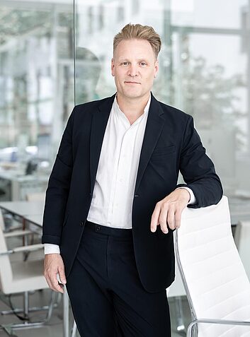 Architekt im dunklen Anzug und weißem Hemd steht in modernem Büro neben einem weißen Stuhl