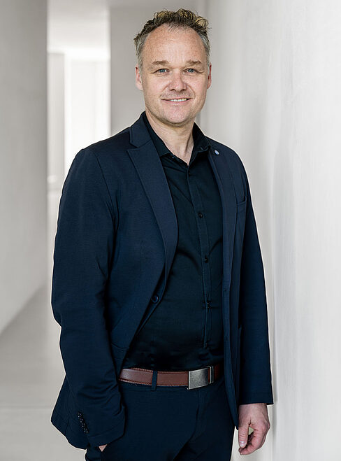 Mann in Anzug steht vor einer Wand in einem hellen Raum.