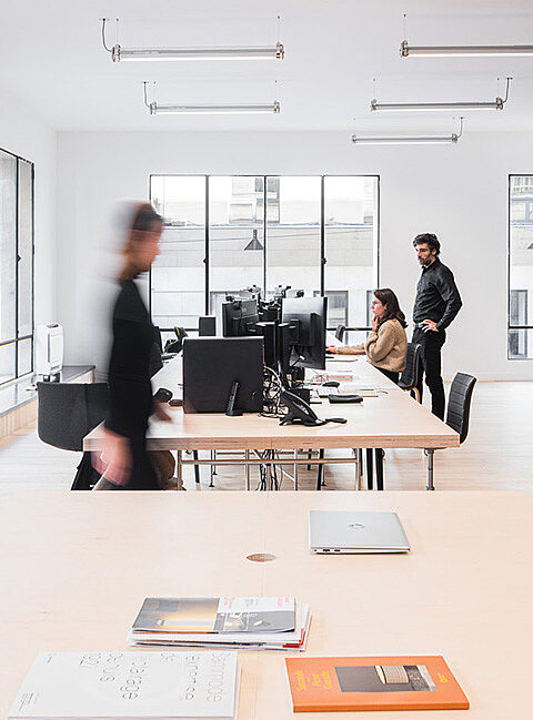 Büro mit mehreren Arbeitsplätzen, Computern, Tischen und Stühlen. Eine Person bewegt sich im Raum.