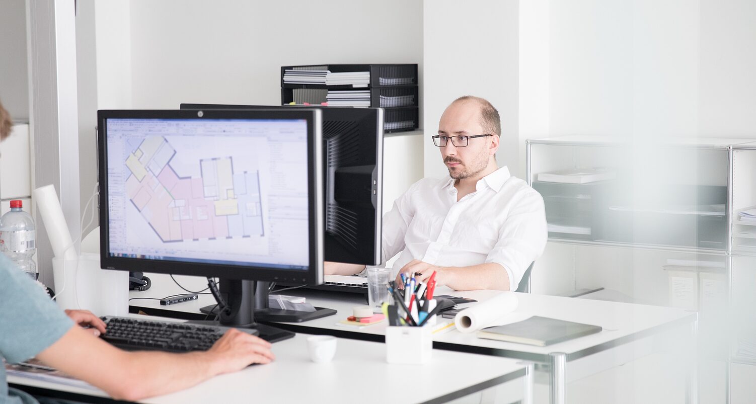 Zwei Personen in einem Büro, eine schaut auf einen Computerbildschirm mit Grundriss, Schreibtisch mit Büromaterial.