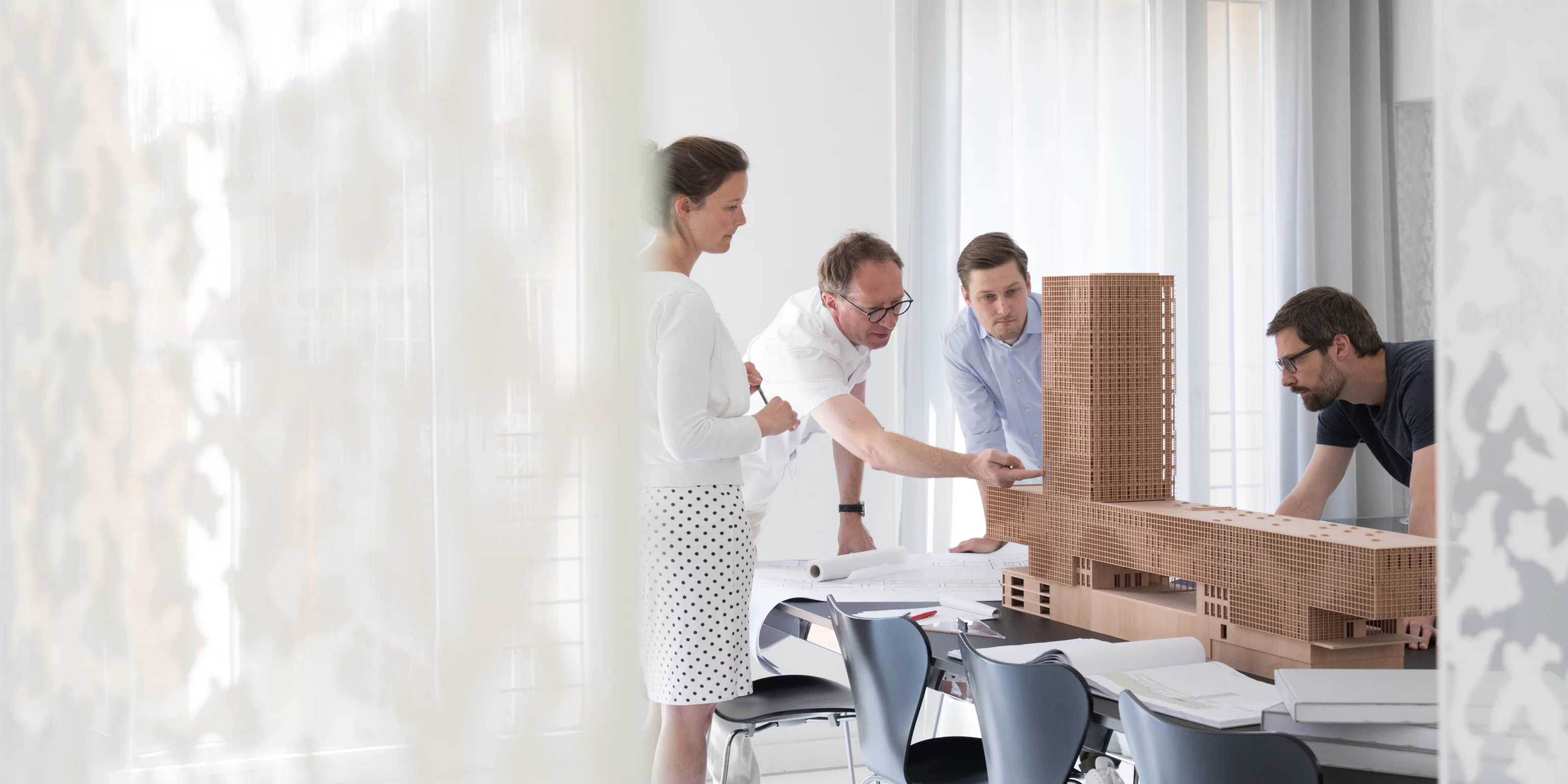 Vier Personen besprechen einen Architekturentwurf an einem Tisch mit einem Modell und mehreren Bauplänen.