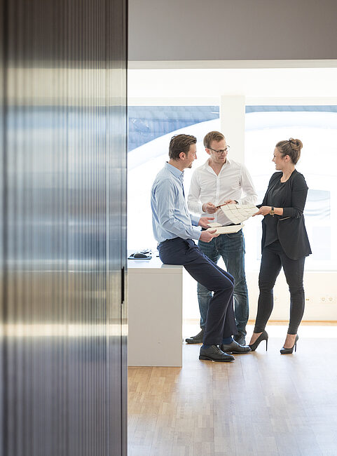 Drei Personen stehen in einem modernen Büro, diskutieren und halten Dokumente. Holzverkleidung im Hintergrund.