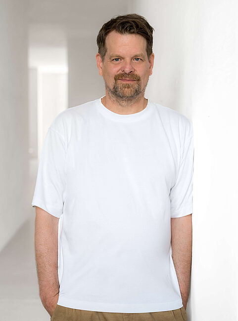 Middle-aged man in white T-shirt leaning on a white wall, looking at the camera
