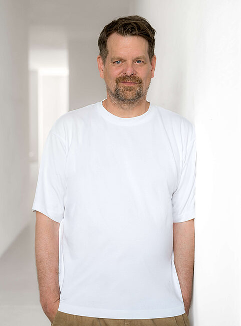 Mann mittleren Alters im weißen T-Shirt lehnt an weißer Wand und blickt in die Kamera