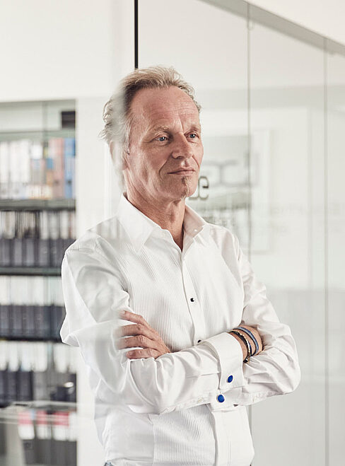 Ein Mann in einem weißen Hemd steht mit verschränkten Armen vor einer Glaswand in einem Büro. Bücherregale im Hintergrund.