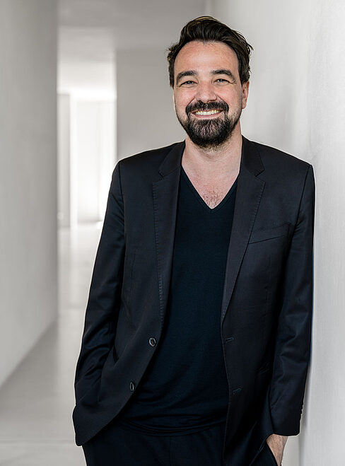 Mann in schwarzem Anzug steht in einem hellen, minimalistischen Raum mit weißen Wänden.