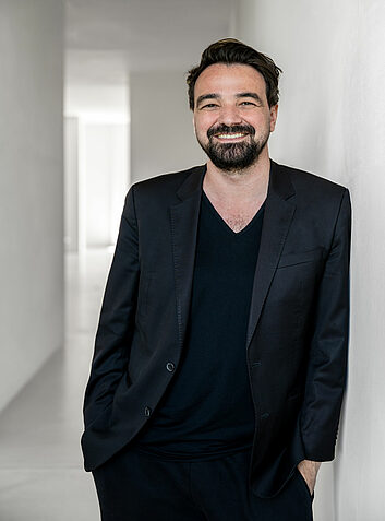 Smiling man in black jacket leaning casually against light-coloured wall