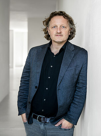 Thoughtful man with curly hair and dark blue jacket leans against a white wall