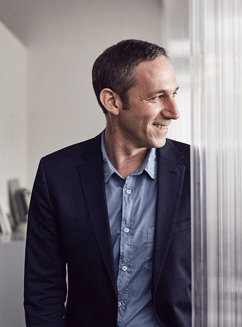 Ein Mann in einem Anzug steht neben einer transparenten Wand. Er lächelt und schaut zur Seite.