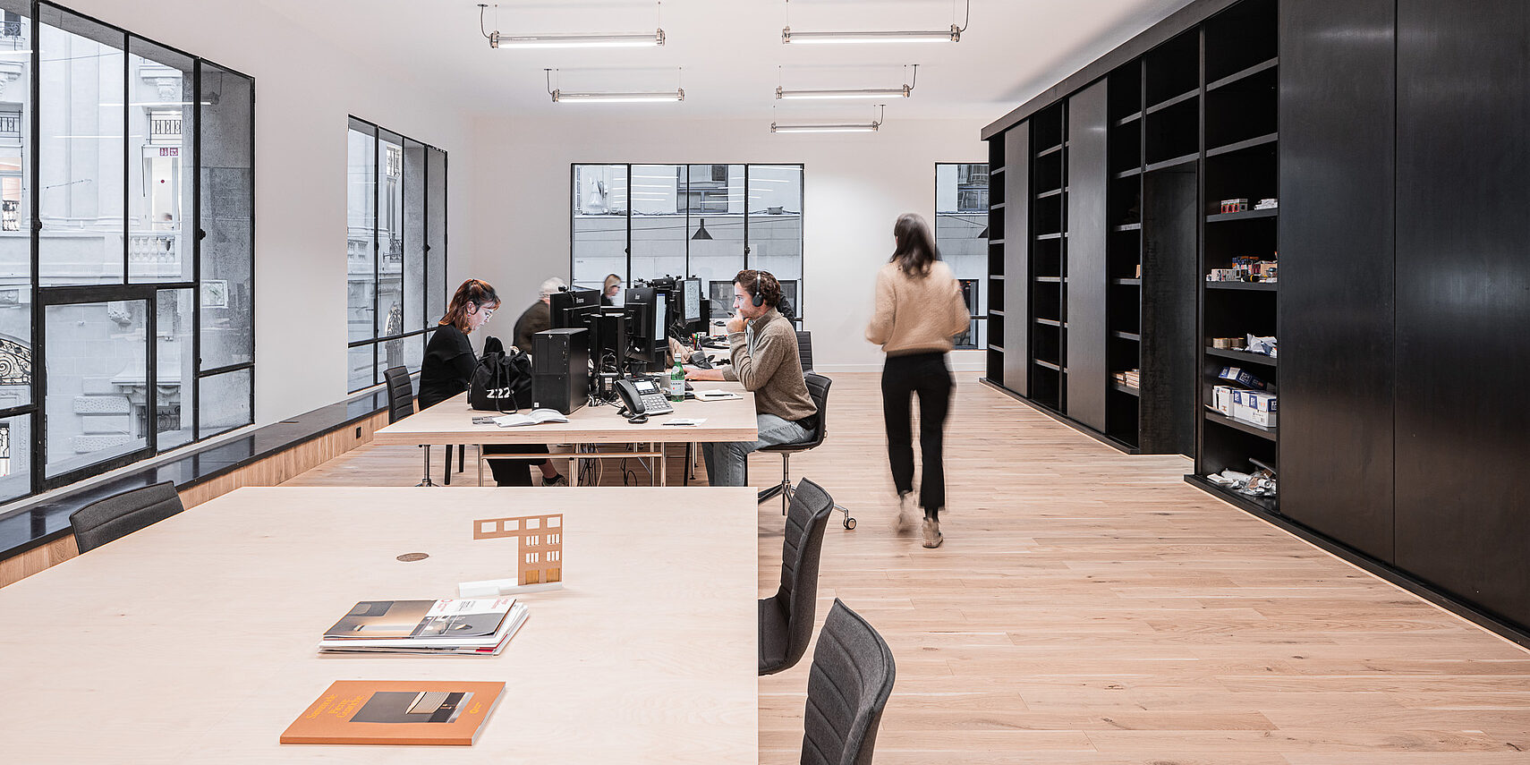 Büro mit Tischen, Computern, Stühlen und großen Fenstern. Eine Person steht, eine andere sitzt. Regale an der Wand.