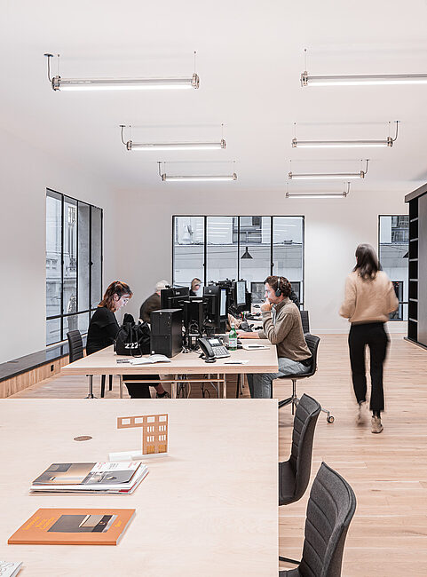 Büro mit mehreren Arbeitsplätzen, Computern, Tischen und Stühlen. Eine Person bewegt sich im Raum.