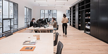 Büro mit mehreren Arbeitsplätzen, Computern, Tischen und Stühlen. Eine Person bewegt sich im Raum.