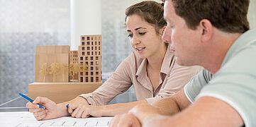 Zwei Personen diskutieren über Pläne, während Modelle von Gebäuden im Hintergrund stehen.
