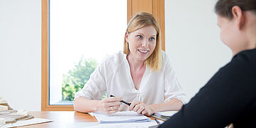 Zwei Frauen sitzen an einem Tisch, eine hält einen Stift, auf dem Tisch liegen Notizblätter und ein Modell.